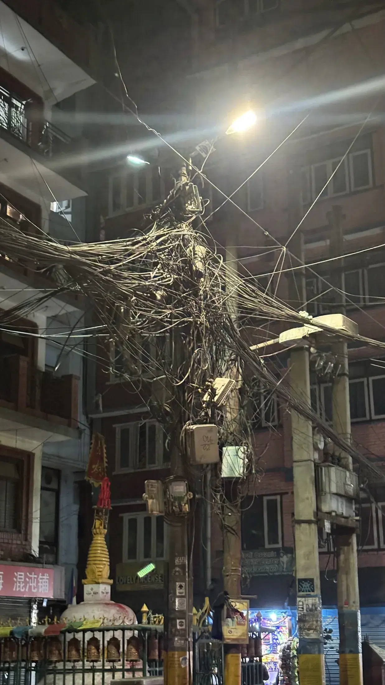 A small street shrine and prayer wheels beneath overhead cables in Kathmandu at night