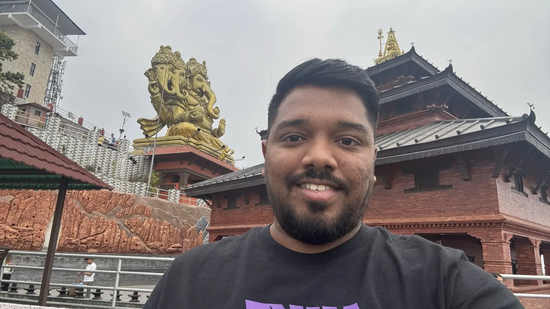 Manish at Pumdikot with a golden Ganesha sculpture and red-roofed temple buildings behind him