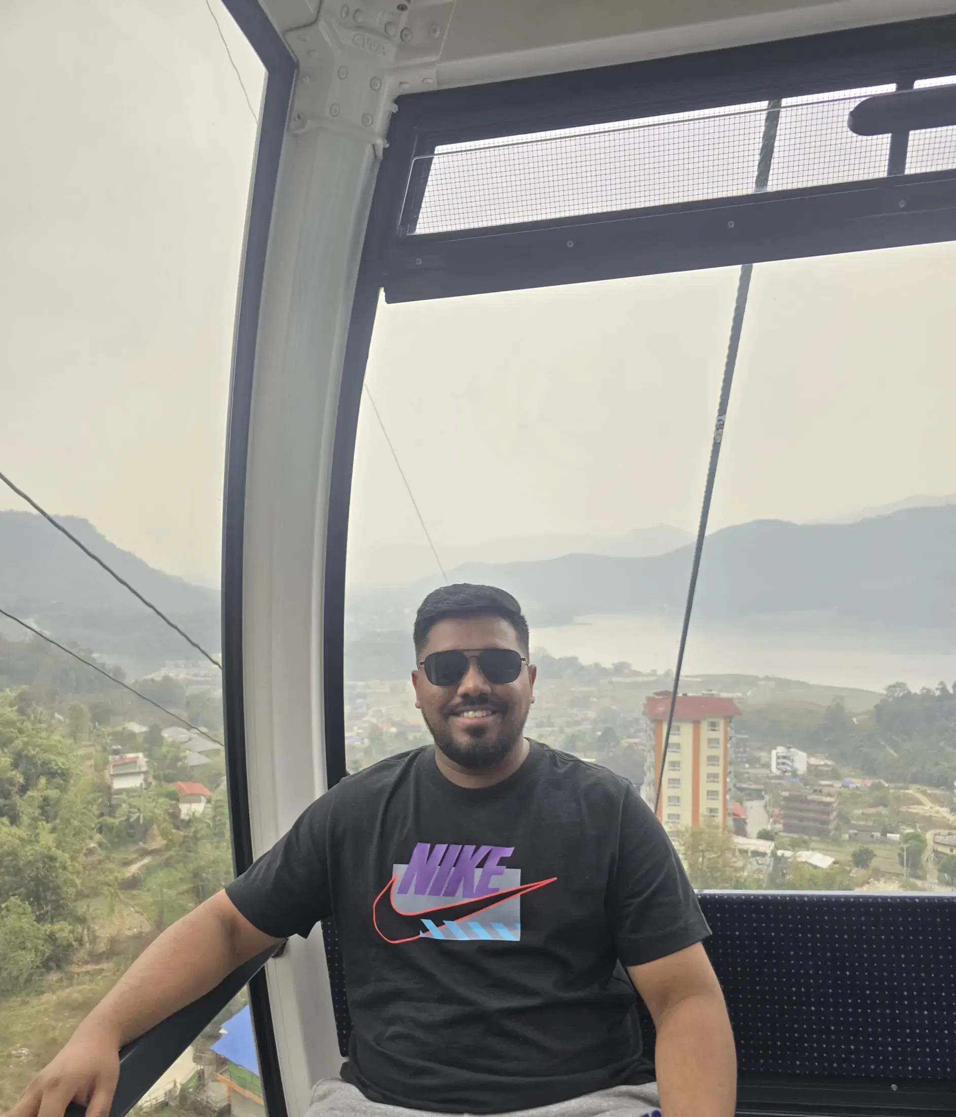 Manish inside a cable-car cabin with Pokhara and Phewa Lake behind him
